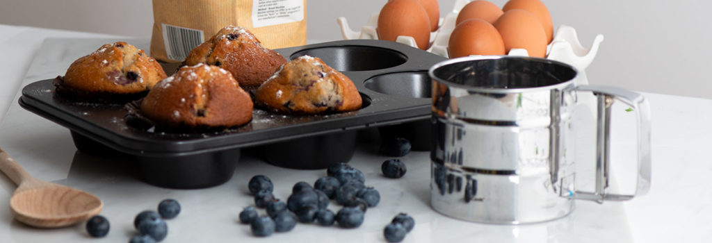 ProCook Muffin Tin and Flour Sifter alongside baked blueberry muffins and a scattering of fresh blueberries